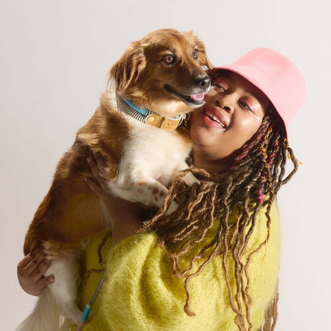 Person with dreadlocks holding a dog against a plain background