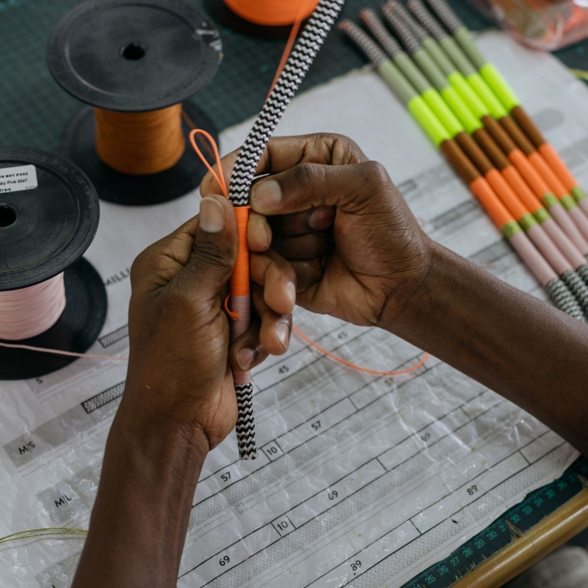 Close-up of a person's hands wrapping an orange cord around a black-and-white striped rope, with spools of thread and finished striped cords arranged on a worktable.